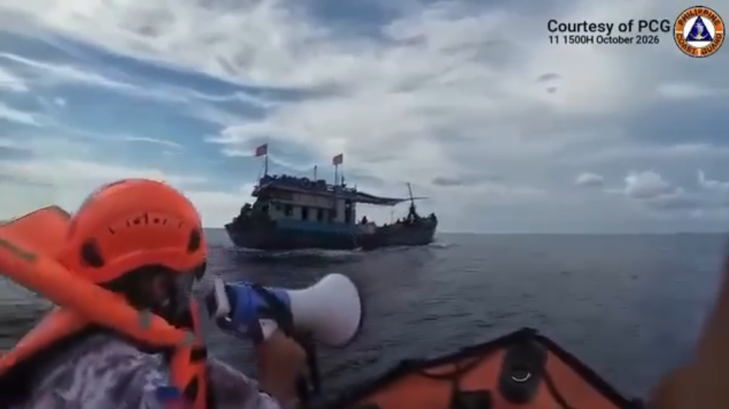Coast guard official in orange helmet holding a megaphone on a boat near another vessel at sea.